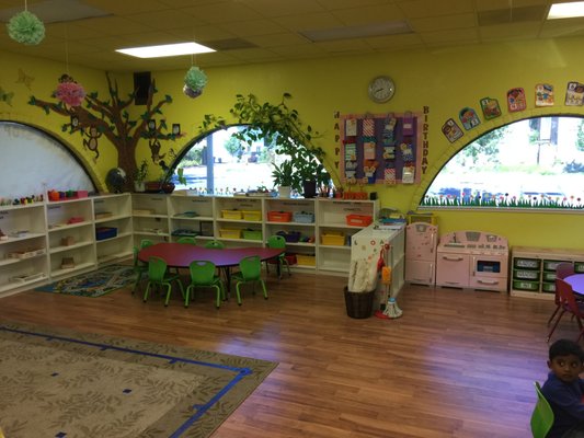 Bright, inviting preschool classroom with colorful walls, shelves of toys, and green tables. A child plays in the corner, enhancing the lively atmosphere.