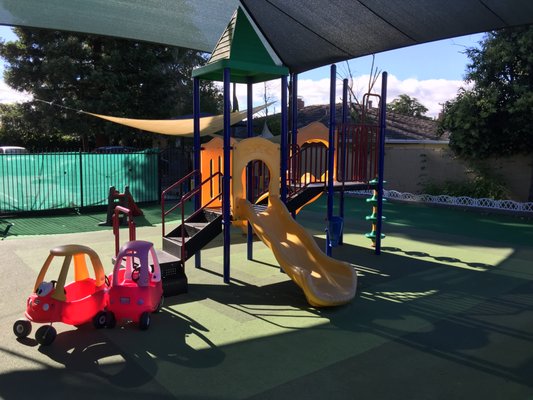 Colorful playground featuring a yellow slide, climbing structure, and red ride-on cars, set in a shaded area, ideal for children's play.