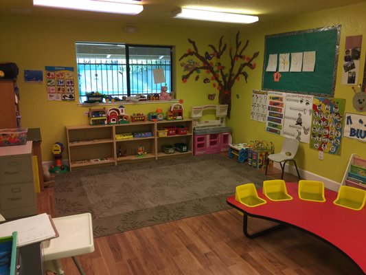 Bright preschool classroom with colorful toy storage, a nature-themed mural, and a red activity table with yellow chairs. Ideal for early learning.