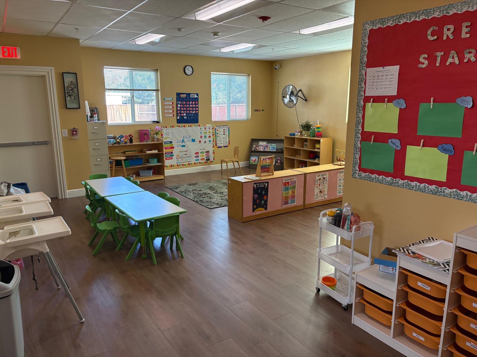 Bright, colorful classroom with green tables and chairs, educational posters, and storage units. Designed for young children's learning and play.