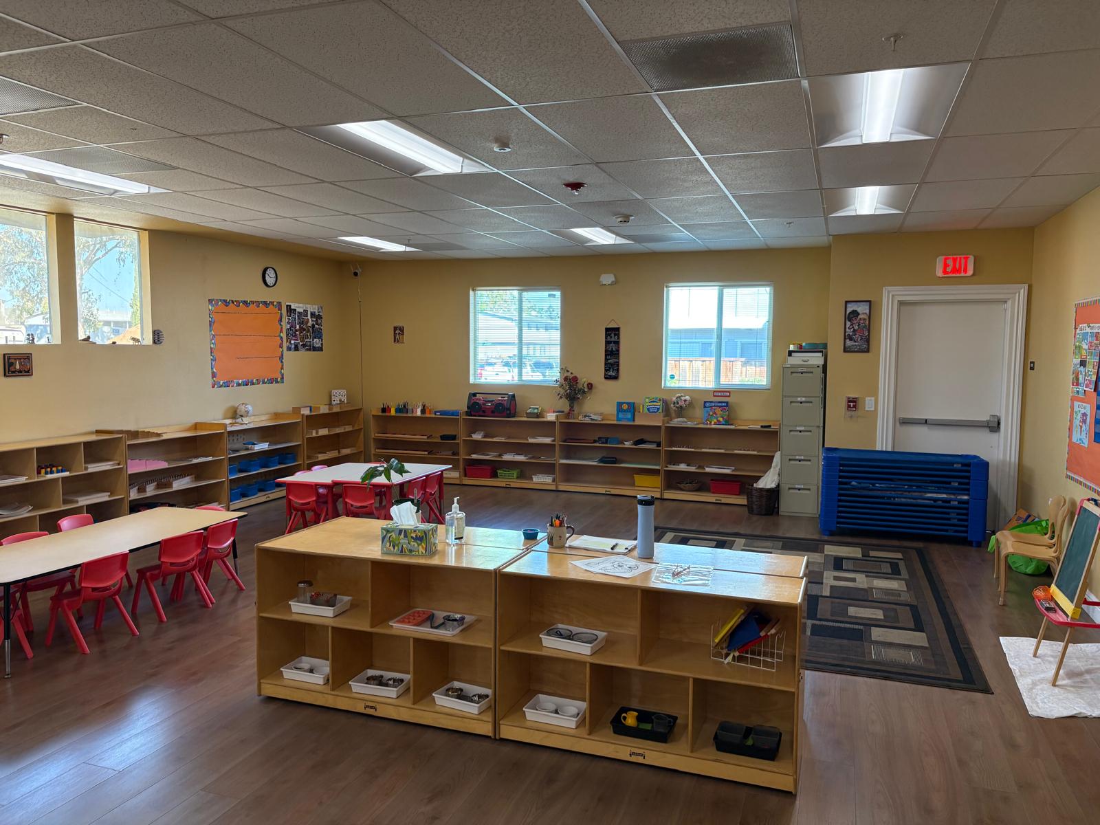 Bright classroom featuring wooden shelves filled with supplies, red chairs, and large windows letting in natural light, promoting a learning environment.