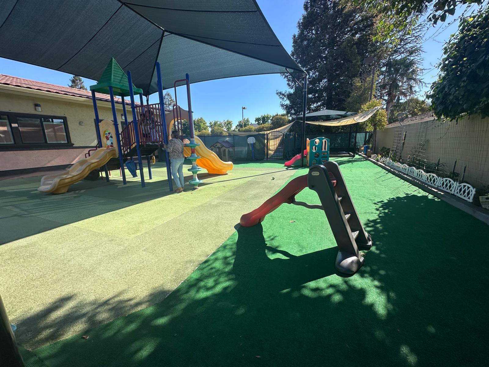 Playground with colorful slides and climbing structure under a shaded canopy. A child interacts with play equipment in a safe, outdoor setting.