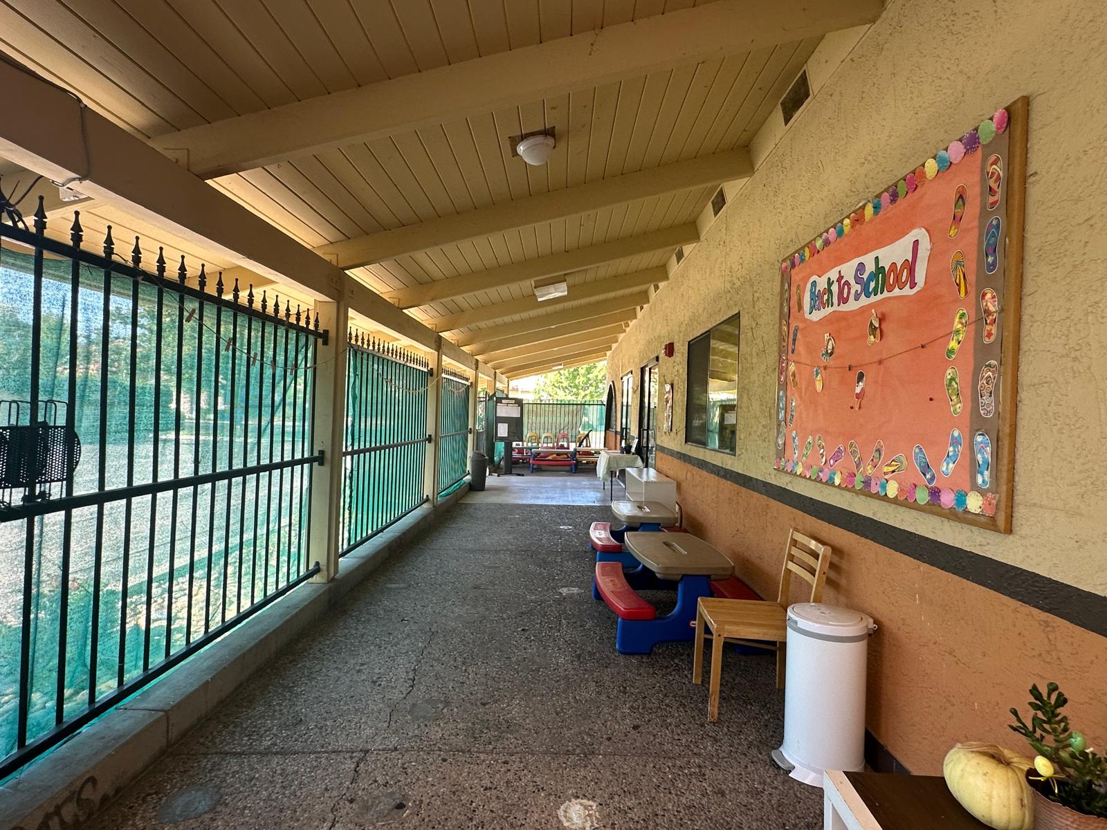 Covered walkway leading to a colorful "Back to School" bulletin board, with tables and chairs on one side, enhancing a vibrant educational environment.